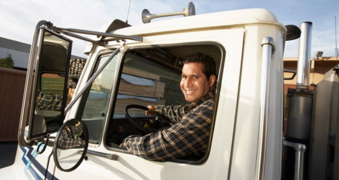 A trucker sits in the cab of a semi truck and smiles towards the camera.