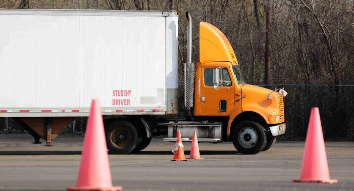 An orange semi truck with "Student Driver" on the side is parked in a lot surrounded by safety cones.
