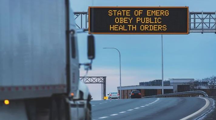 A semi truck drives up to a freeway sign that reads "State of emerg obey publick health orders."