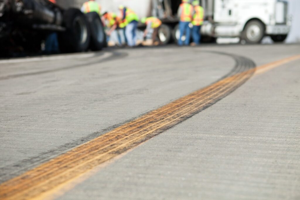 A closeup of tire skid marks on the road with a large group of workers in safety vests in the background.
