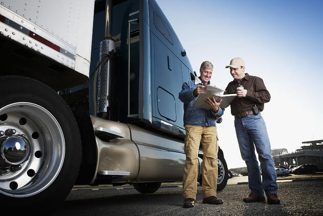 Two truckers stand outside the cab of a semi truck and review documents in a clipboard.