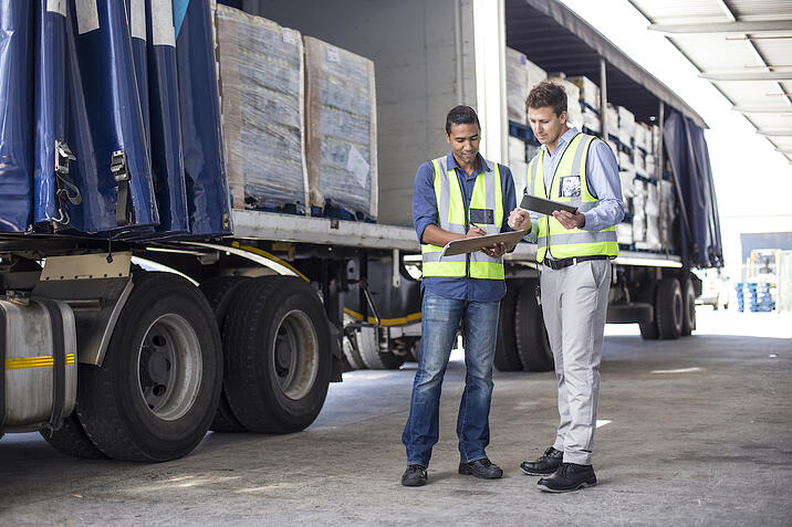 Two workers in safety vests stand next to a semi truck and review documents