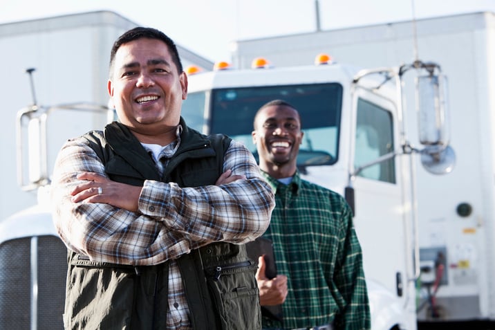 Two semi truck drivers stand in front of a commercial truck and smile to the camera