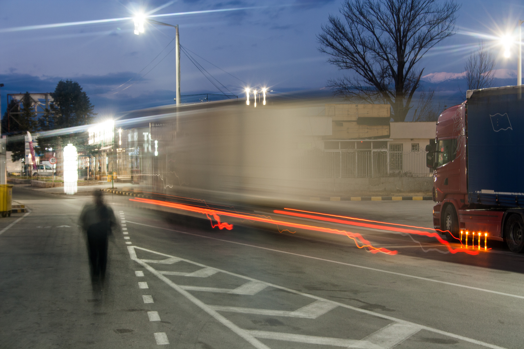 A semi truck driving down a city street at night, while a pedestrian walks alongside it.